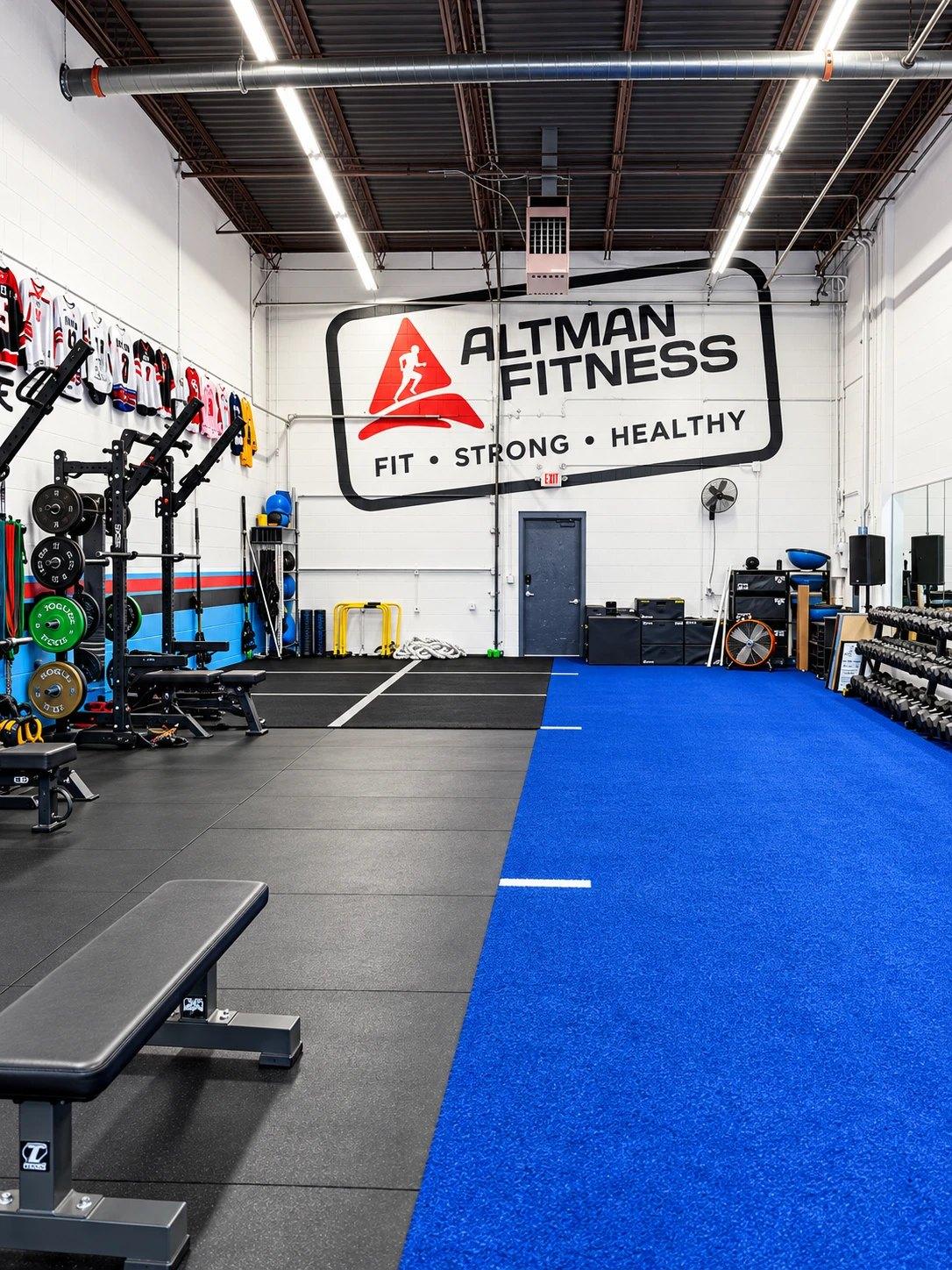 The Altman Fitness training floor in Edina: squat racks and benches on rubber flooring beside a blue turf lane, under the Fit · Strong · Healthy wall sign