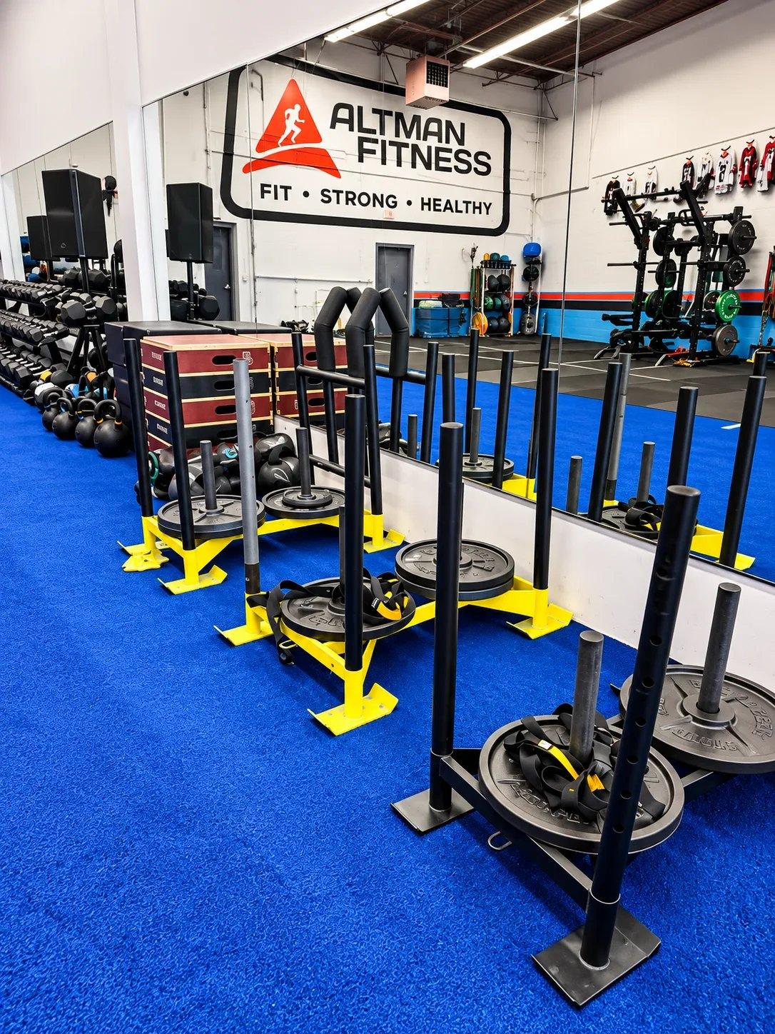 Push sleds loaded with plates and suspension straps lined up on the turf at Altman Fitness, with dumbbells, kettlebells and plyo boxes behind